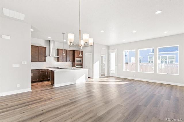 kitchen featuring wall chimney range hood, light hardwood / wood-style flooring, appliances with stainless steel finishes, a kitchen island with sink, and hanging light fixtures