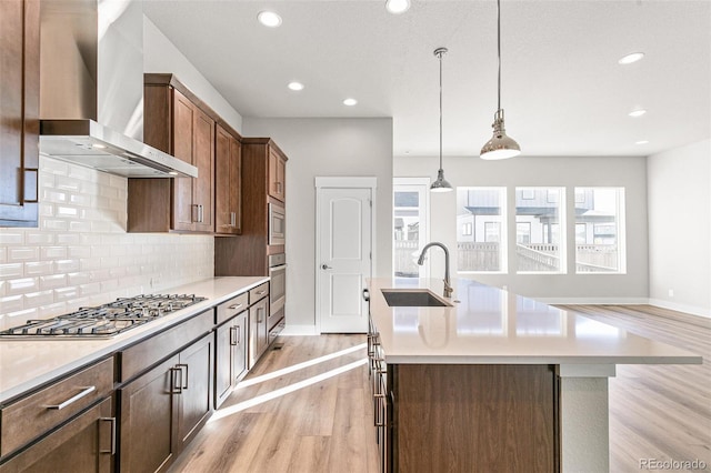 kitchen with sink, appliances with stainless steel finishes, an island with sink, wall chimney exhaust hood, and light wood-type flooring