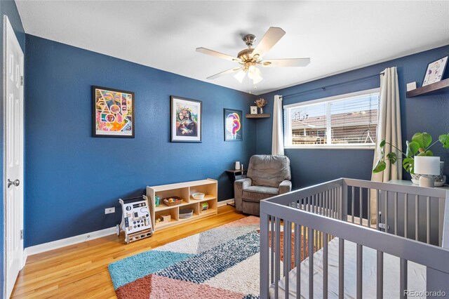 bedroom with a ceiling fan, a nursery area, baseboards, and wood finished floors