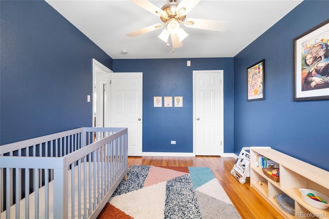 bedroom with a crib, wood finished floors, a ceiling fan, and baseboards