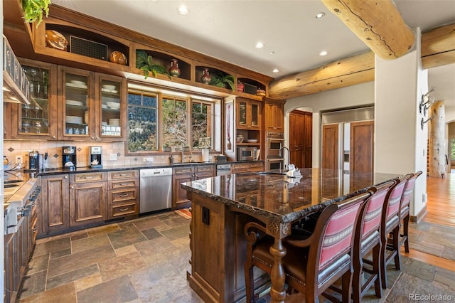 kitchen featuring a kitchen island with sink, dark stone counters, sink, decorative backsplash, and appliances with stainless steel finishes