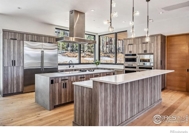 kitchen with appliances with stainless steel finishes, decorative light fixtures, island exhaust hood, a center island, and light hardwood / wood-style floors