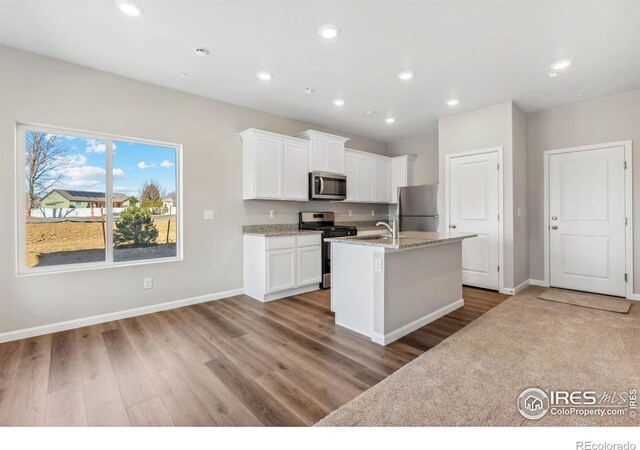 kitchen with a kitchen island with sink, light stone countertops, white cabinetry, stainless steel appliances, and light hardwood / wood-style floors