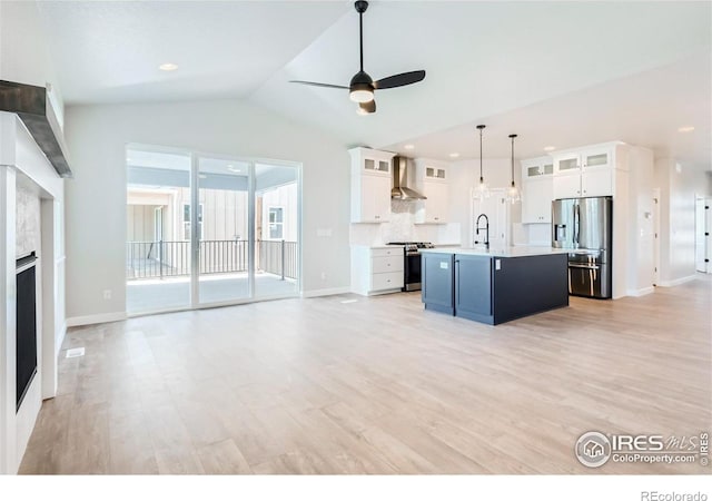 kitchen with wall chimney exhaust hood, hanging light fixtures, stainless steel appliances, a center island with sink, and light hardwood / wood-style floors