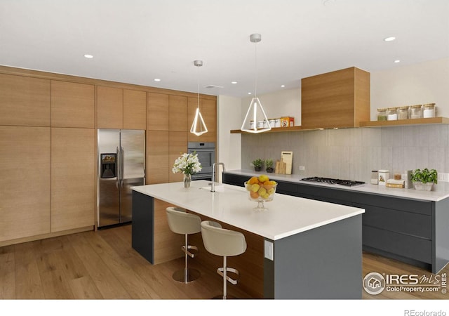 kitchen with stainless steel appliances, backsplash, pendant lighting, a center island with sink, and light wood-type flooring