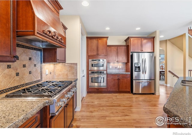 kitchen featuring light stone counters, decorative backsplash, light hardwood / wood-style flooring, appliances with stainless steel finishes, and premium range hood