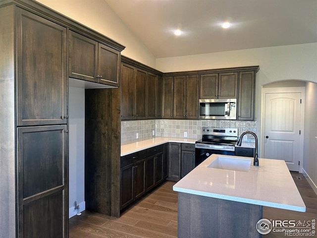 kitchen with dark hardwood / wood-style flooring, lofted ceiling, an island with sink, and appliances with stainless steel finishes