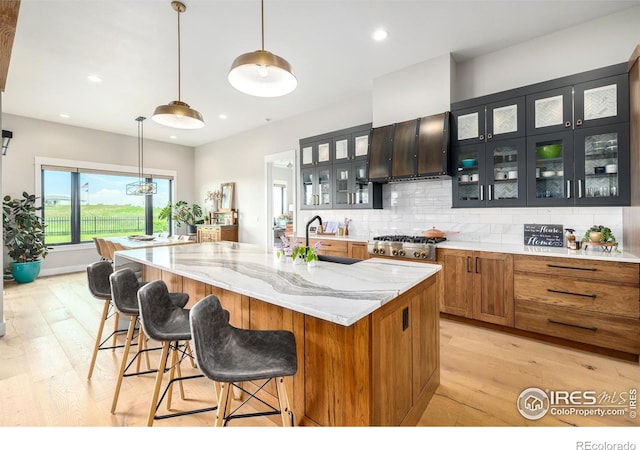 kitchen with wall chimney range hood, light hardwood / wood-style flooring, tasteful backsplash, light stone countertops, and an island with sink