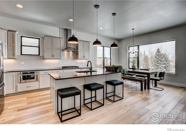 kitchen with appliances with stainless steel finishes, sink, hanging light fixtures, a kitchen island with sink, and wall chimney range hood