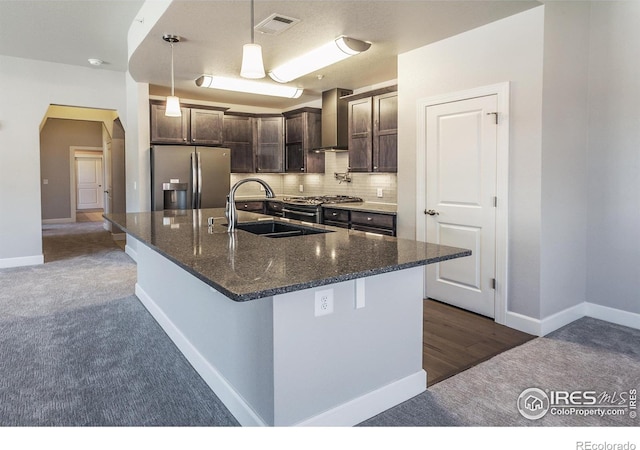 kitchen with a kitchen island with sink, dark stone counters, wall chimney range hood, stainless steel fridge, and dark brown cabinets