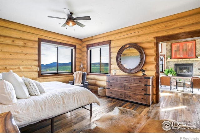 bedroom with hardwood / wood-style flooring, ceiling fan, a stone fireplace, and rustic walls