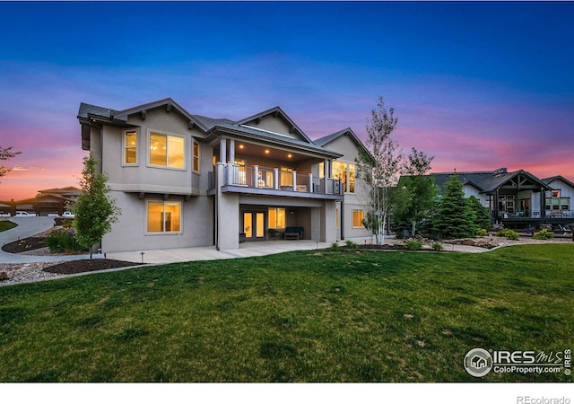 back house at dusk with a lawn, a balcony, and a patio