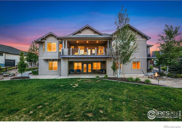 back house at dusk with a lawn, french doors, a balcony, a patio area, and an outdoor fire pit