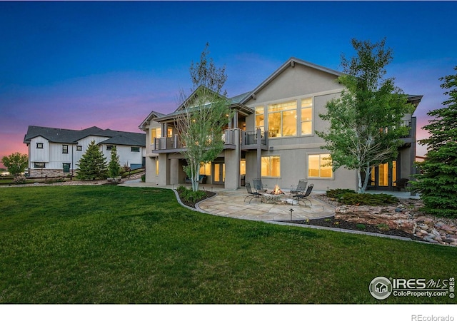 back house at dusk with a patio, a balcony, a fire pit, and a lawn