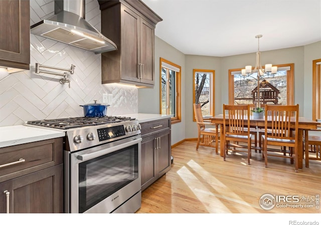 kitchen with wall chimney exhaust hood, light wood-type flooring, tasteful backsplash, an inviting chandelier, and stainless steel gas stove