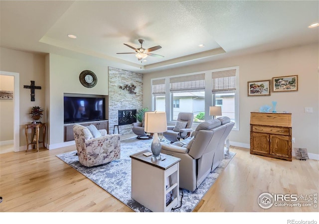 living room with ceiling fan, a stone fireplace, light hardwood / wood-style floors, a textured ceiling, and a tray ceiling