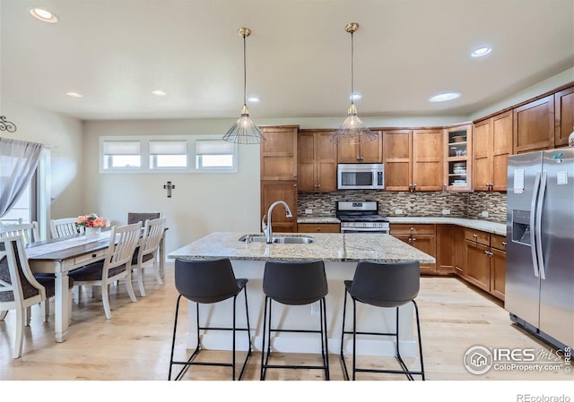 kitchen with light stone counters, stainless steel appliances, a kitchen island with sink, sink, and pendant lighting