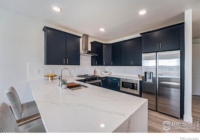 kitchen with kitchen peninsula, stainless steel appliances, sink, wall chimney range hood, and light hardwood / wood-style floors