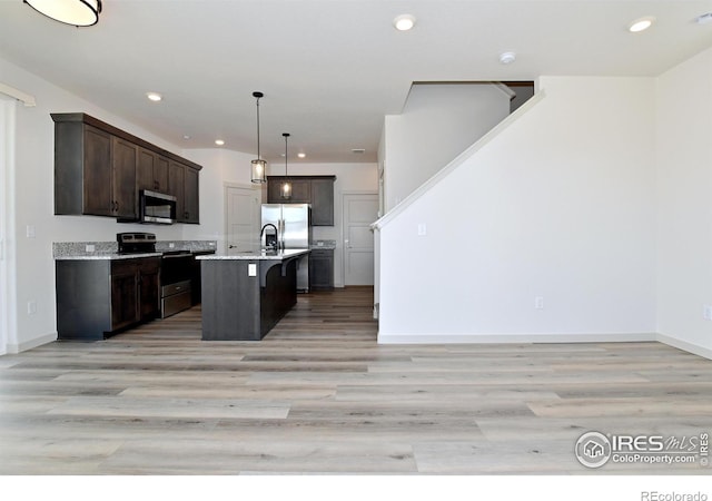 kitchen featuring appliances with stainless steel finishes, decorative light fixtures, a kitchen island with sink, dark brown cabinetry, and light wood-type flooring