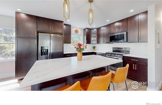 kitchen with a center island, light wood-type flooring, light stone countertops, appliances with stainless steel finishes, and decorative light fixtures