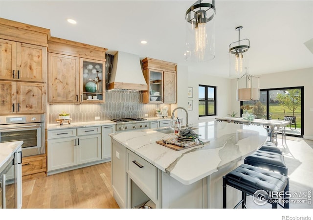 kitchen with sink, hanging light fixtures, custom range hood, a center island with sink, and appliances with stainless steel finishes