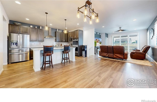 kitchen with appliances with stainless steel finishes, wall chimney exhaust hood, dark brown cabinets, a center island, and a breakfast bar area