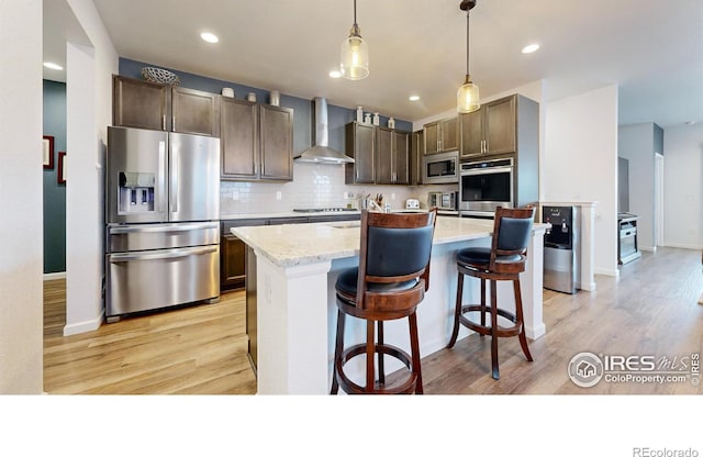 kitchen with wall chimney exhaust hood, light stone counters, pendant lighting, a kitchen island with sink, and appliances with stainless steel finishes