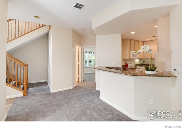 kitchen with white appliances, dark stone counters, light brown cabinetry, light colored carpet, and kitchen peninsula