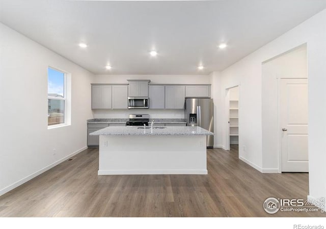 kitchen with stainless steel appliances, gray cabinetry, light hardwood / wood-style floors, light stone countertops, and an island with sink