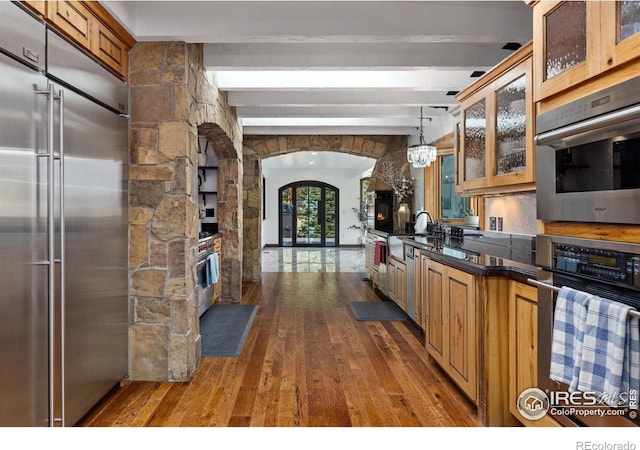 kitchen featuring arched walkways, stainless steel appliances, dark countertops, hanging light fixtures, and glass insert cabinets