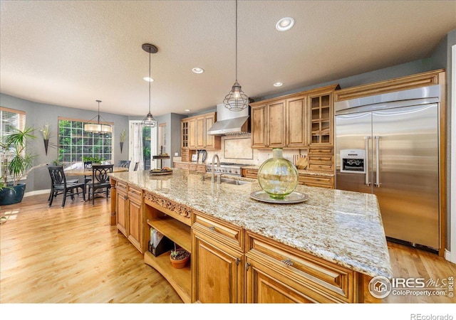 kitchen featuring stainless steel built in refrigerator, wall chimney exhaust hood, an island with sink, decorative light fixtures, and sink