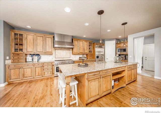 kitchen with a center island with sink, appliances with stainless steel finishes, light stone counters, pendant lighting, and wall chimney range hood