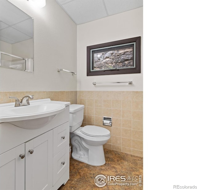 bathroom with a wainscoted wall, tile walls, toilet, vanity, and a drop ceiling