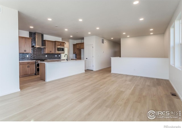 kitchen with light wood-type flooring, appliances with stainless steel finishes, an island with sink, wall chimney range hood, and backsplash