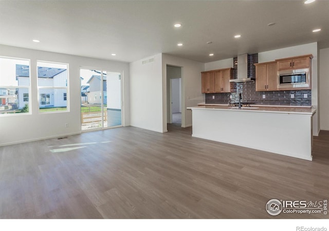 kitchen featuring hardwood / wood-style flooring, a kitchen island with sink, stainless steel microwave, tasteful backsplash, and wall chimney exhaust hood