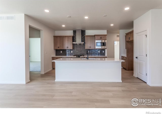 kitchen featuring sink, light wood-type flooring, stainless steel microwave, a kitchen island with sink, and wall chimney range hood