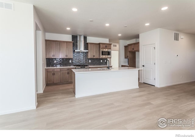 kitchen featuring sink, a kitchen island with sink, stainless steel appliances, wall chimney exhaust hood, and light wood-type flooring