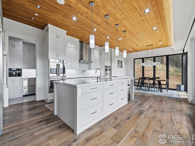 kitchen with dark hardwood / wood-style floors, a kitchen island with sink, backsplash, decorative light fixtures, and wall chimney range hood