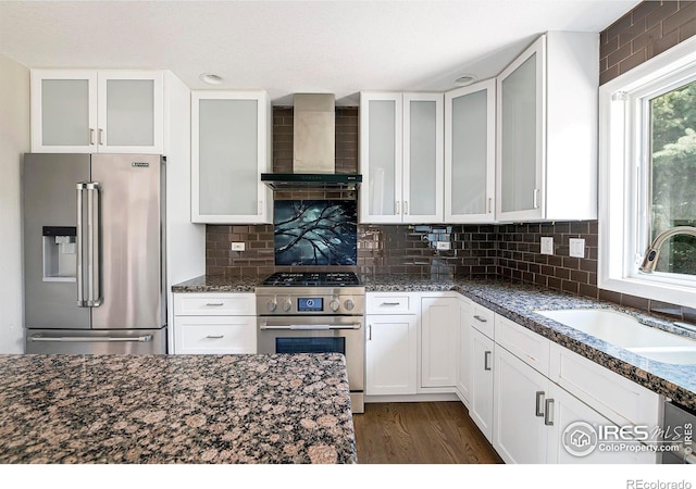 kitchen with sink, wall chimney range hood, dark stone countertops, stainless steel appliances, and white cabinets