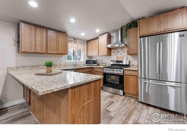 kitchen with stainless steel appliances, light stone counters, kitchen peninsula, wall chimney exhaust hood, and light wood-type flooring