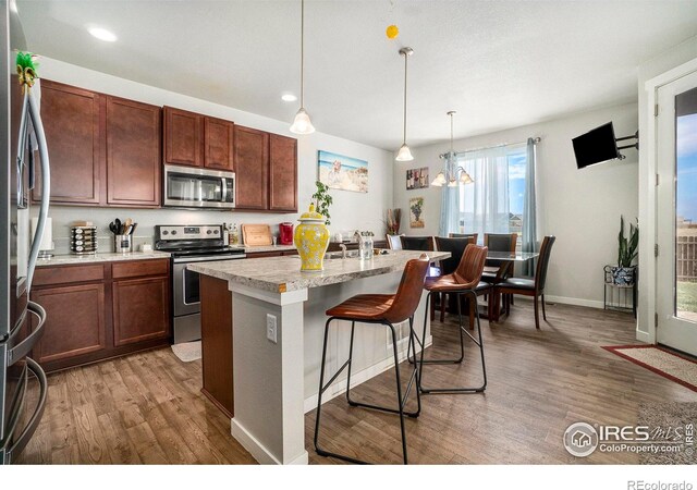 kitchen with appliances with stainless steel finishes, wood-type flooring, decorative light fixtures, and a center island with sink