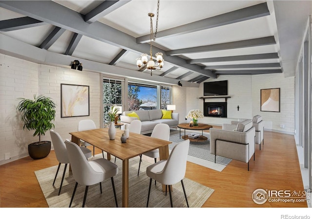 dining room featuring brick wall, lofted ceiling with beams, a chandelier, and a glass covered fireplace