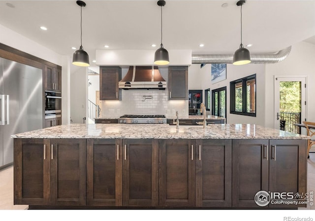 kitchen with wall chimney range hood, backsplash, a kitchen island with sink, appliances with stainless steel finishes, and light stone countertops