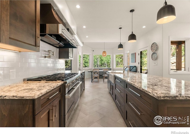 kitchen with light tile patterned flooring, wall chimney exhaust hood, tasteful backsplash, double oven range, and a center island with sink