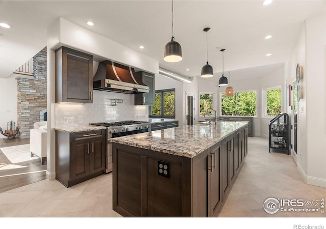 kitchen featuring wall chimney exhaust hood, gas stove, light stone countertops, a kitchen island with sink, and light tile patterned floors