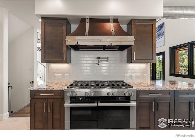 kitchen with backsplash, wall chimney exhaust hood, stainless steel gas stove, light stone counters, and light tile patterned floors