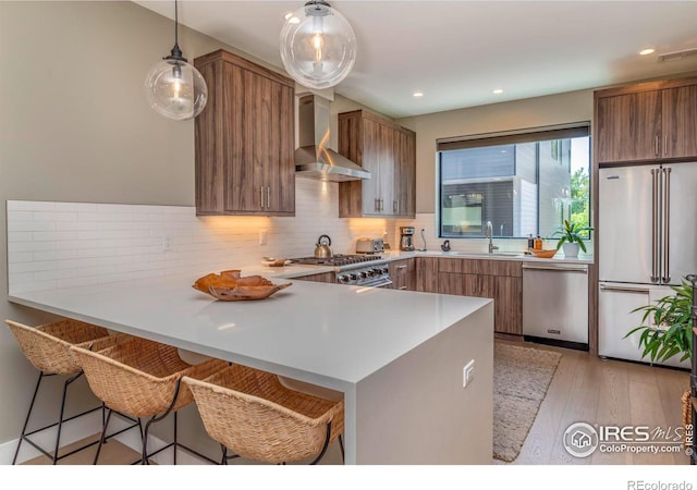 kitchen featuring a peninsula, a sink, high quality appliances, wall chimney range hood, and brown cabinetry