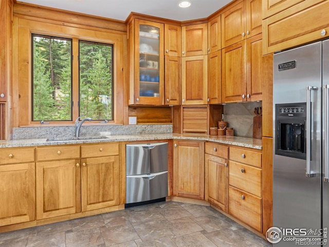 kitchen featuring high end fridge, a sink, dishwashing machine, light stone countertops, and glass insert cabinets