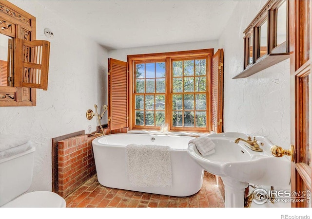 bathroom featuring a soaking tub, toilet, a textured wall, and brick floor