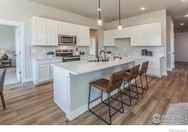 kitchen with stainless steel appliances, white cabinetry, sink, wood-type flooring, and a kitchen island with sink